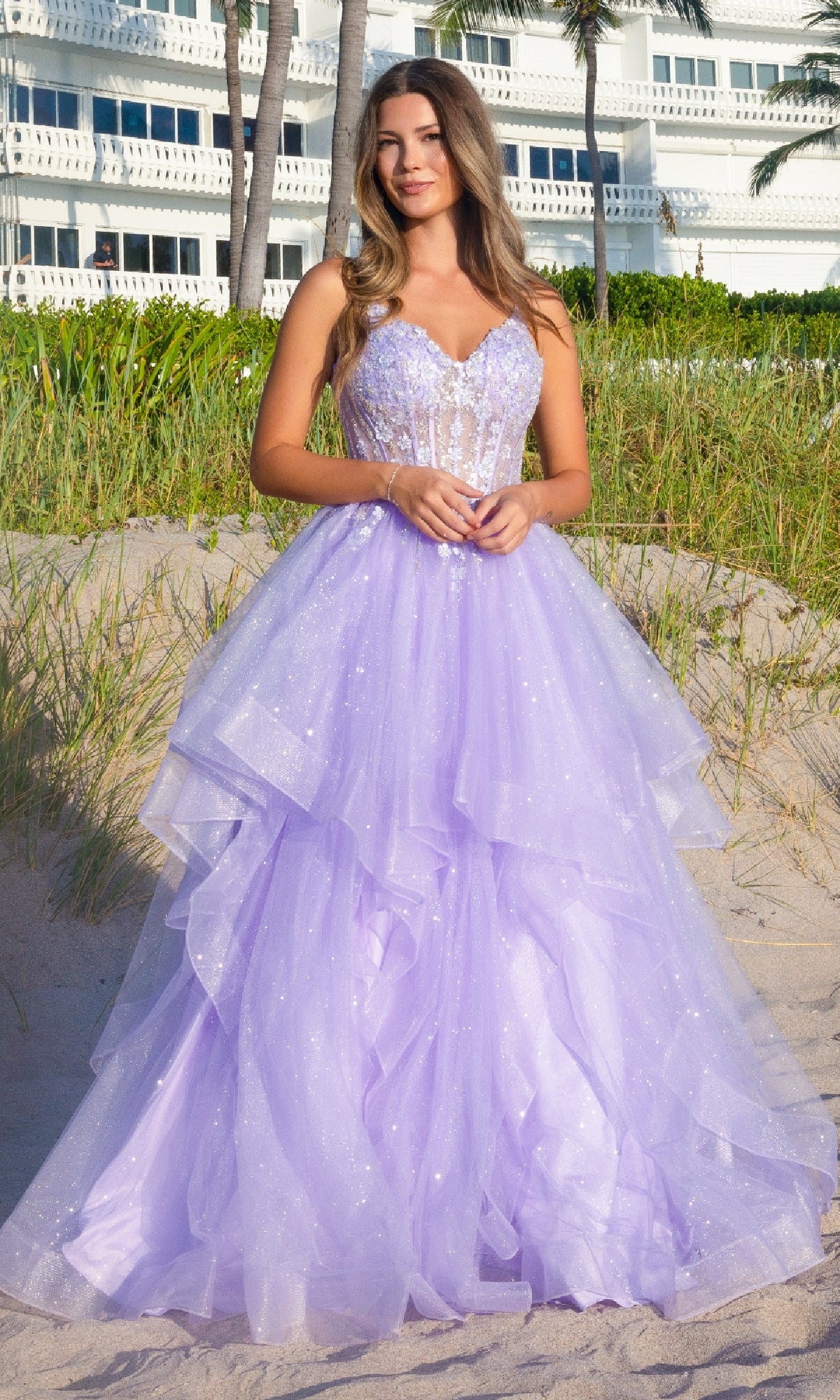 A woman stands on sand before grass and a white building, wearing the Amelia Couture 5068—a long glitter prom ball gown with an ethereal lavender layered tulle skirt and sequined bodice.